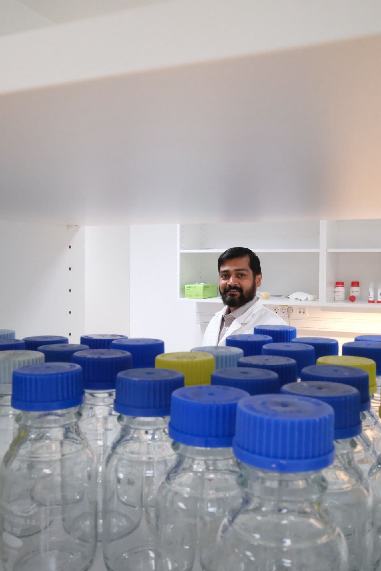 Researcher in a lab coat behind rows of laboratory bottles